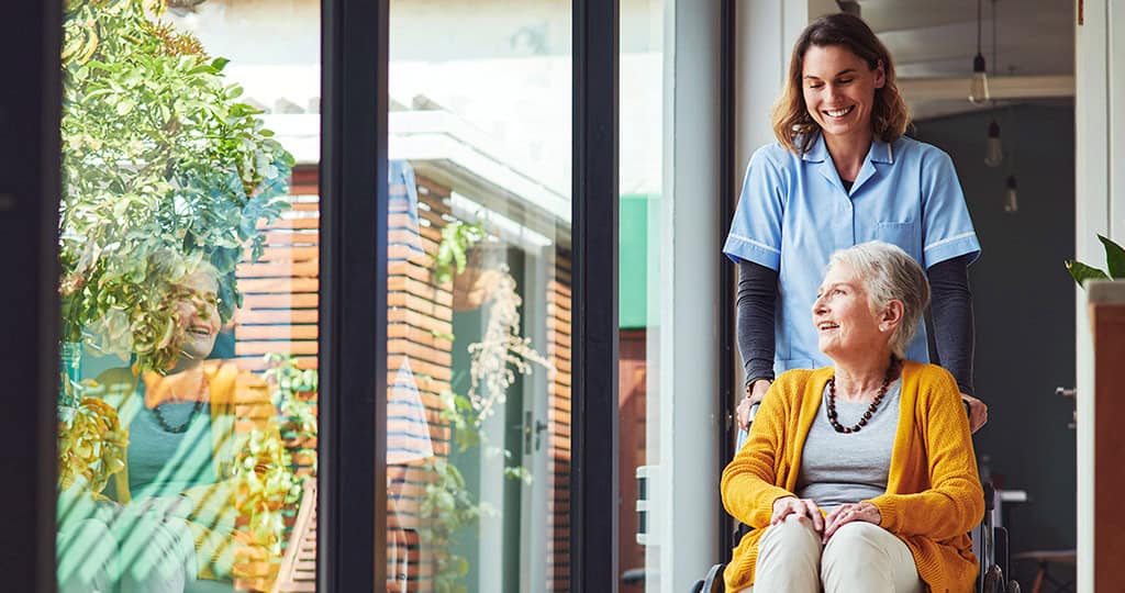 Young female caregiver pushing elderly woman in a wheelchair down hallway in retirement home