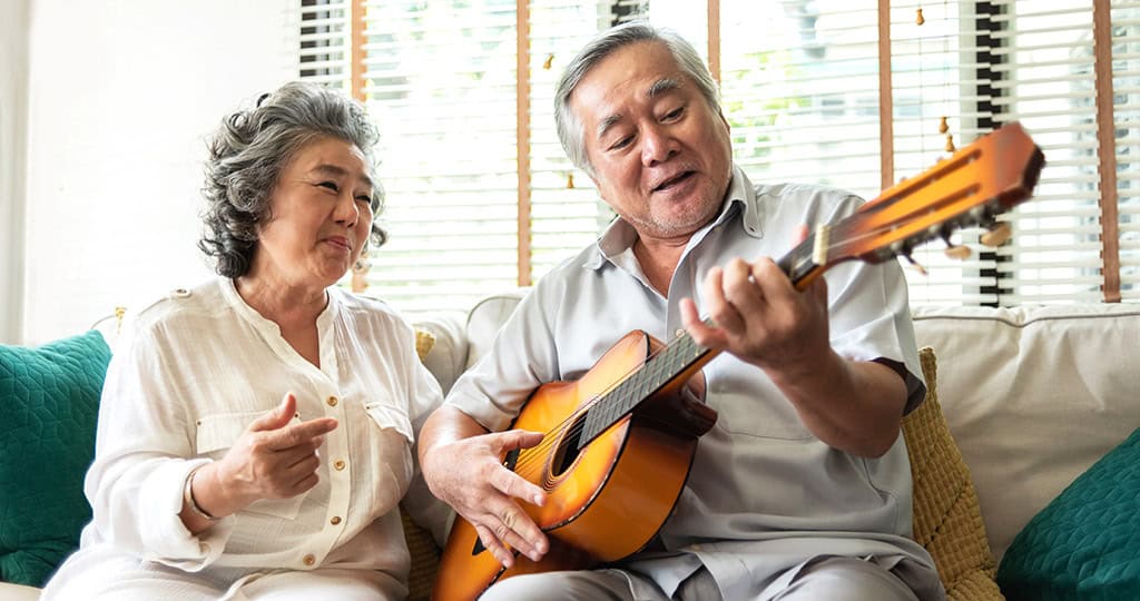 Senior man playing guitar joined by wife singing while sitting on couch