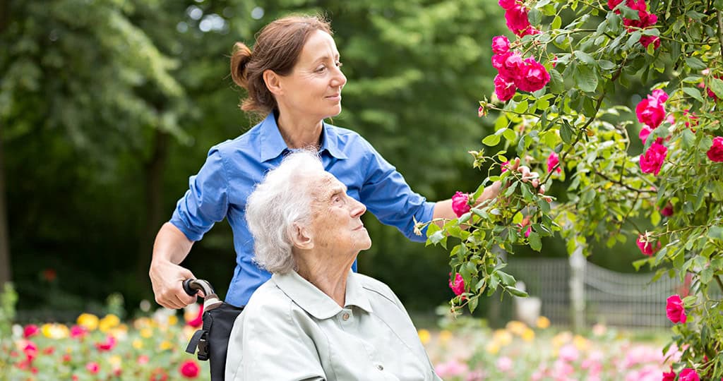 Female caregiver pushing happy woman in wheelchair through garden in the outdoors