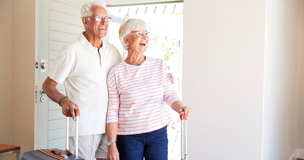 Senior couple looking around at hotel room with smiles on their faces and holding their rolling suitcases