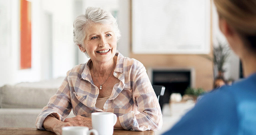 Smiling senior woman holding mug sitting at dining table with caregiver