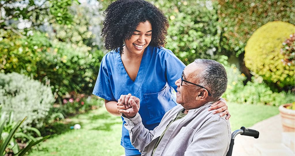 Caregiver from agency smiling and holding hand of senior man in wheelchair