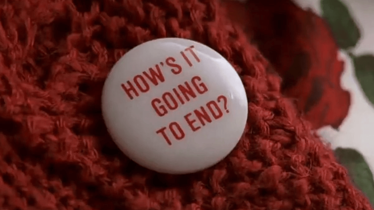 A white pin with red writing is affixed to a red knit sweater. The writing on the pin reads, "How's it going to end?"