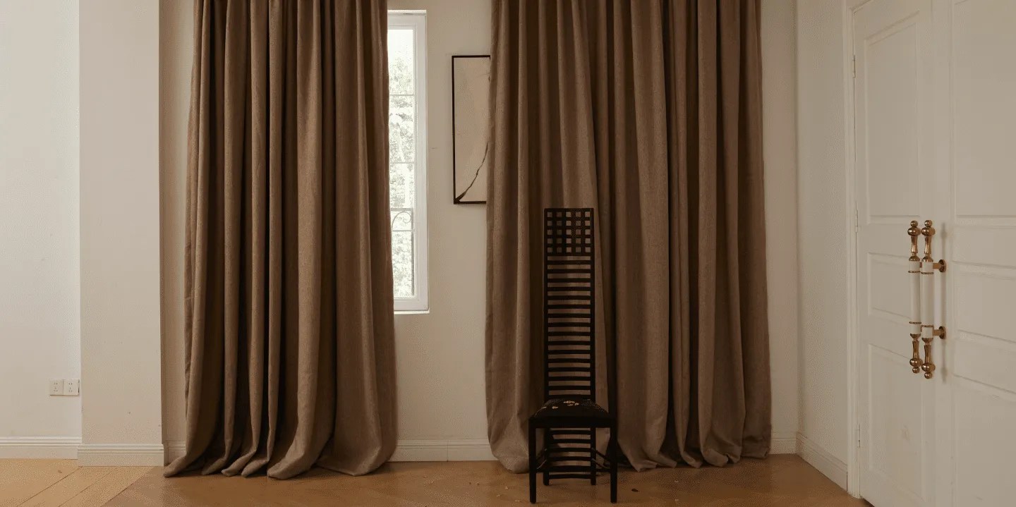 Single wooden chair placed in front of long brown curtains in a minimal room.
