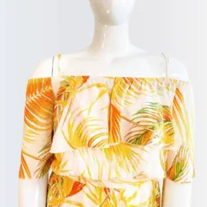 Maniquí luciendo un romper corto con estampado tropical en tonos amarillo, naranja y verde. Diseño con hombros descubiertos, volantes en el escote y tirantes delgados. Silueta suelta y fresca ideal para climas cálidos y días soleados.