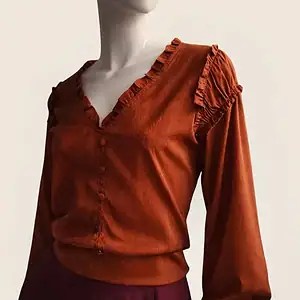 Blusa de color terracota con escote en V y volantes en el contorno, botones forrados y mangas con frunces, sobre maniquí con fondo beige suave.