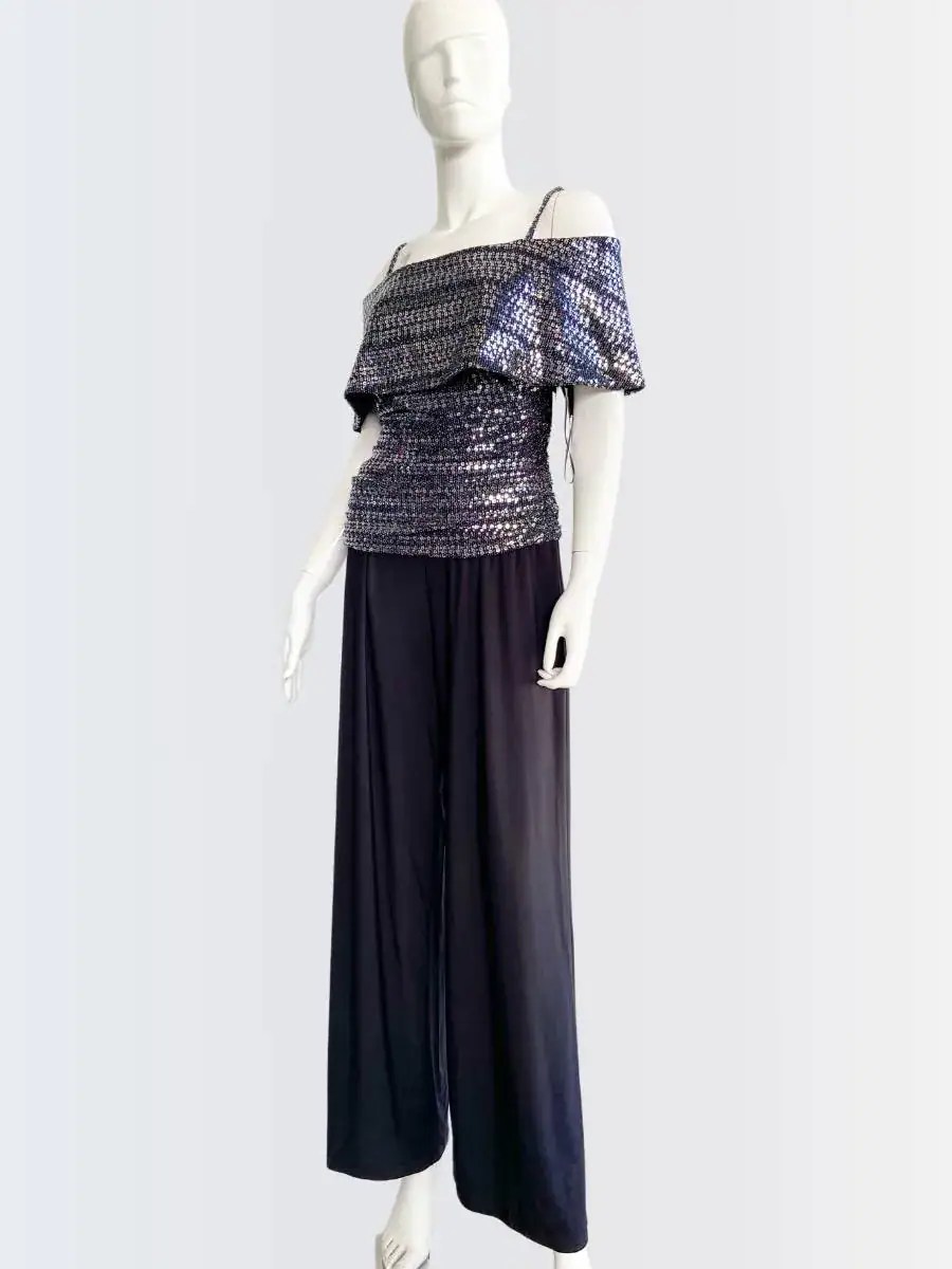 Maniquí vistiendo un jumpsuit de pantalón largo negro con top brillante de lentejuelas en tonos plata y azul, hombros descubiertos y tirantes finos. Diseño elegante y moderno, ideal para ocasiones nocturnas y eventos especiales.