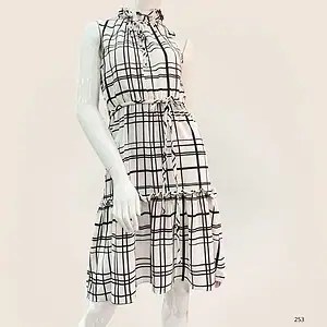 Vestido sin mangas blanco con estampado de cuadros negros, cuello alto con volantes, cinturón ajustable en la cintura y falda con volantes, presentado en maniquí con fondo beige.