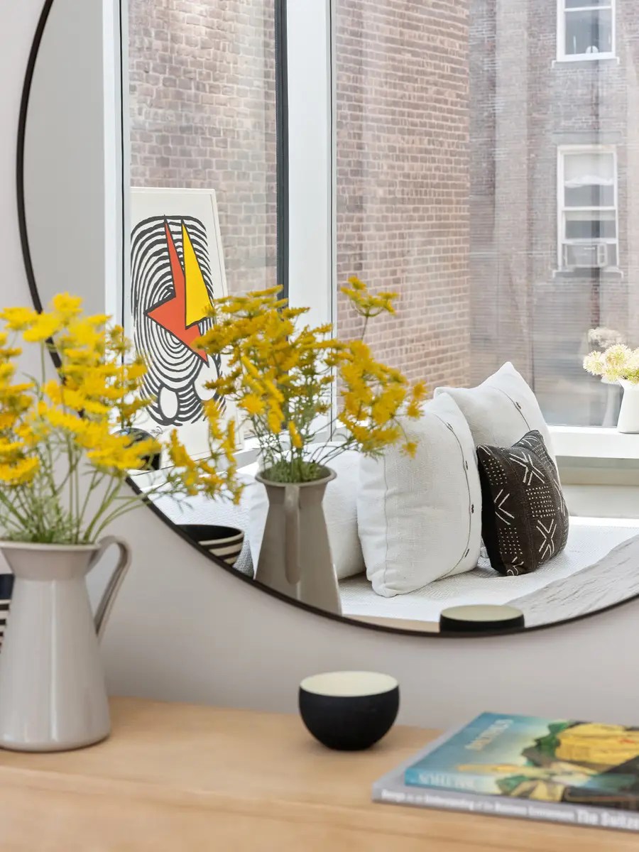 Round mirror reflecting a window seat with linen and mudcloth pillows, framed by yellow wildflowers on a wood console