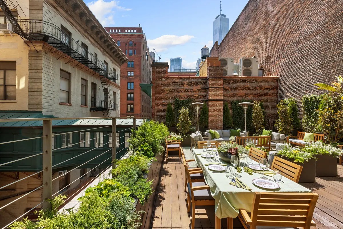 Rooftop dining terrace set for a meal, framed by brick chimneys, potted greenery, and the Manhattan skyline