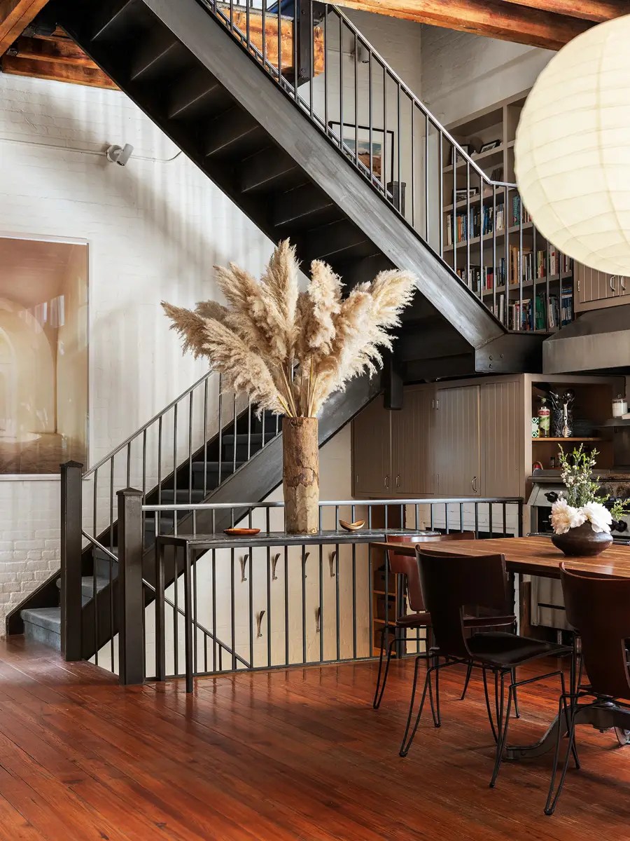 Loft stairwell with pampas grass in a ceramic vase, a black steel staircase, and a mezzanine library above a dining table