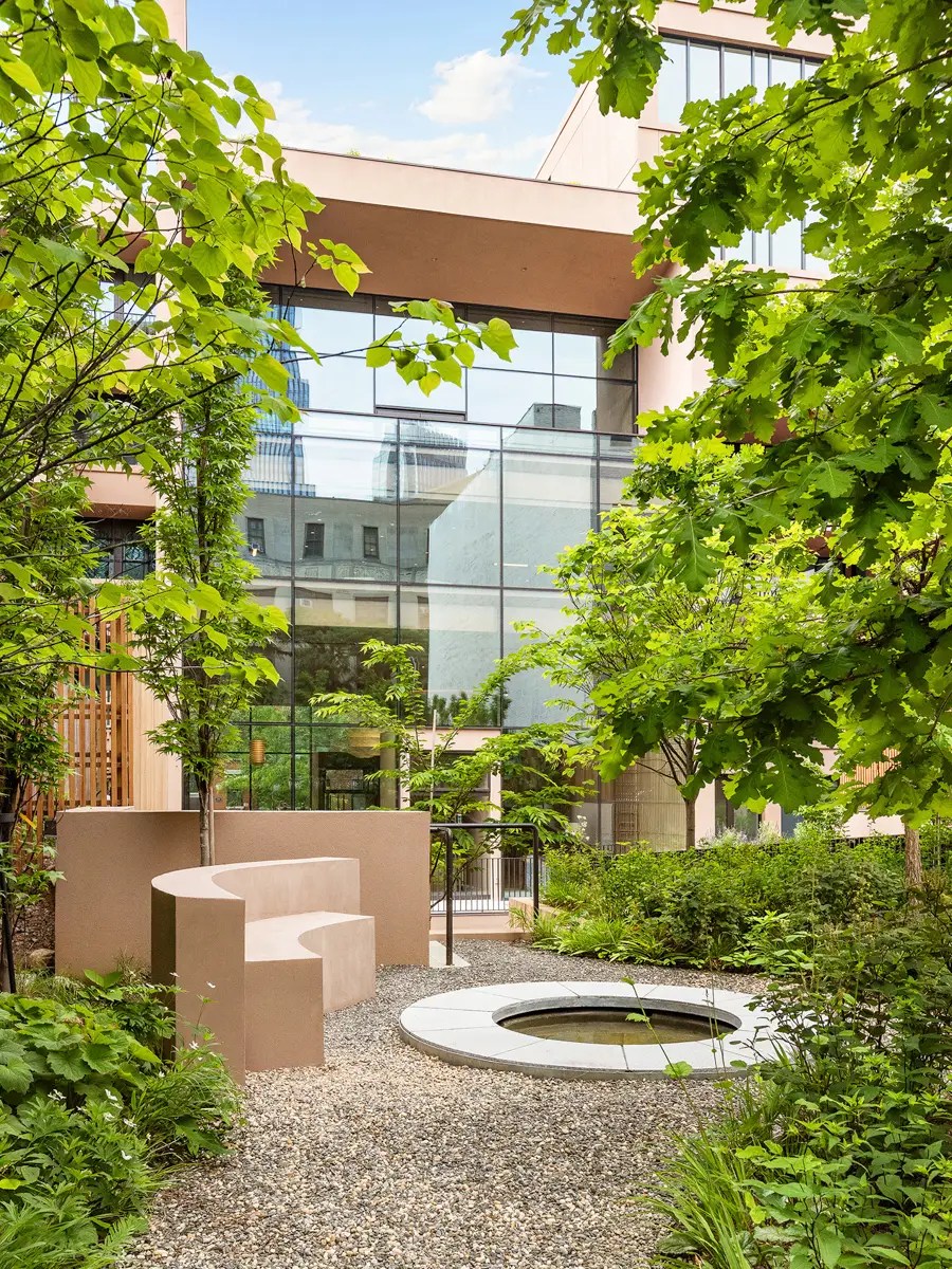 Landscaped courtyard with a sculptural curved concrete bench beside a round reflecting pool, framed by a glass facade and maple trees
