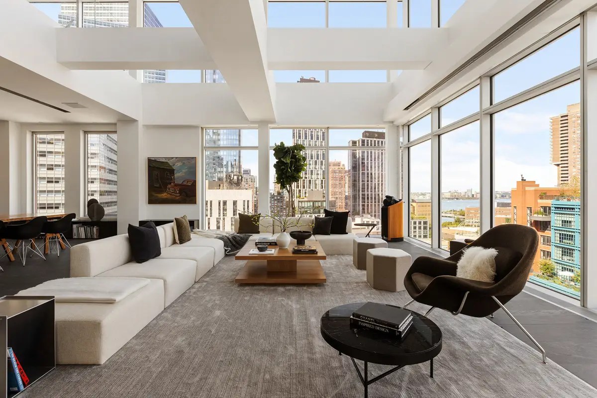 Double-height living room with floor-to-ceiling windows overlooking the Hudson River, a white sectional, and a black leather lounge chair