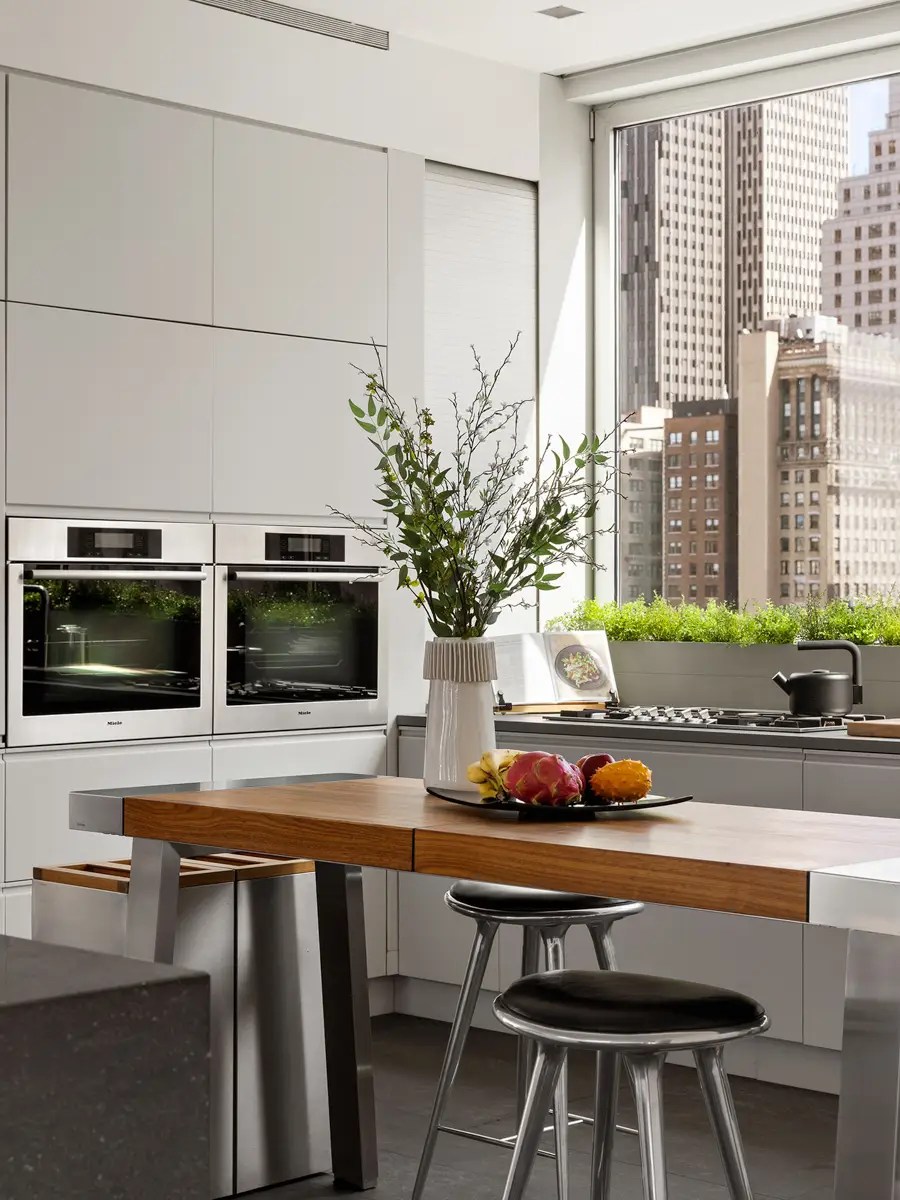 Sleek white kitchen with stainless double ovens, a walnut dining peninsula, and floor-to-ceiling city views