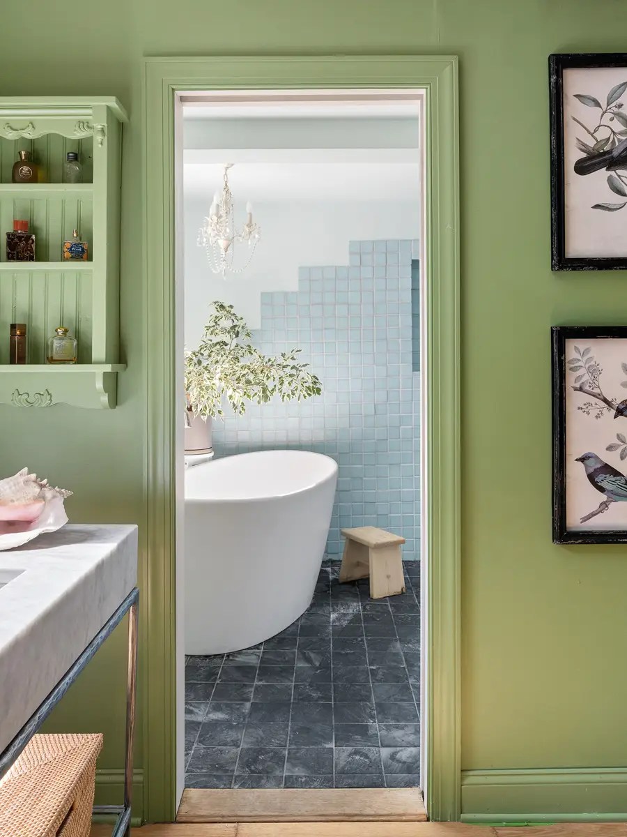 Sage-green hallway framing a doorway view of a freestanding tub against pale blue zellige tile