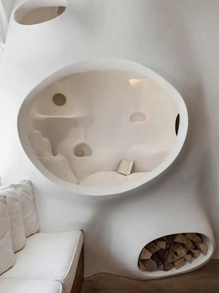Sculptural white plaster reading nook with an organic oval cutout, built-in cubbies, and a curved firewood niche below