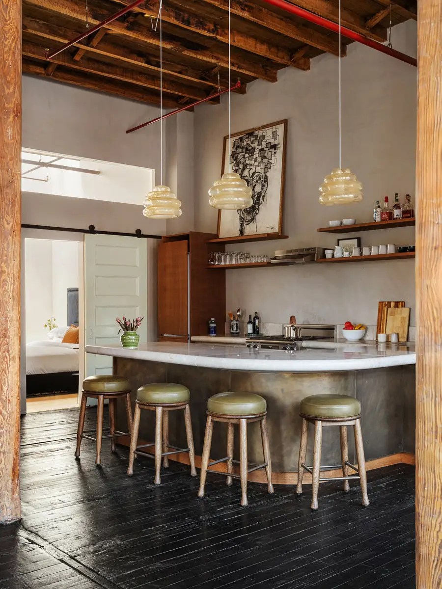 Curved metal kitchen bar with amber glass pendant lights, olive leather stools, and open wood shelving