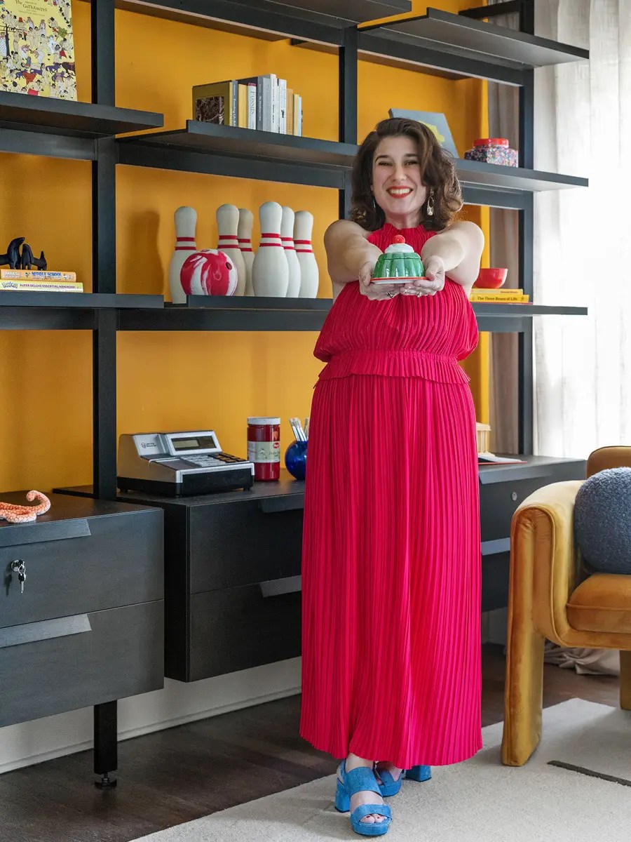 Woman in a red pleated dress holding a green bundt cake in front of a mustard-yellow wall with black shelving