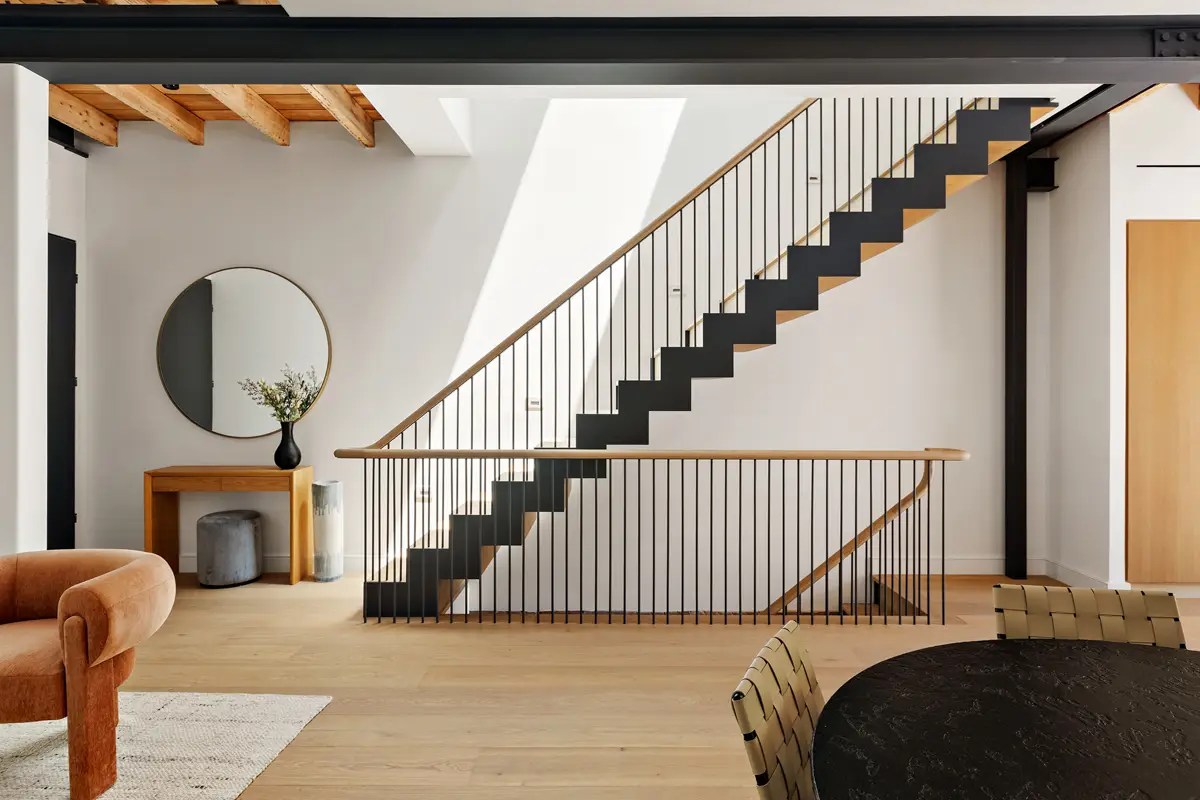 Black steel staircase with wood treads in a white loft, with a round mirror and orange velvet chair nearby