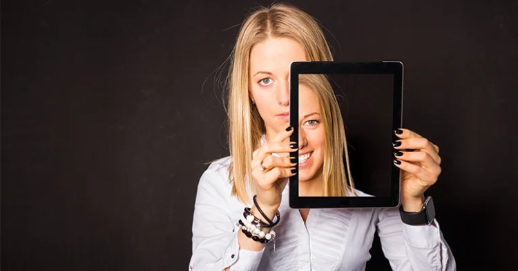 OnlineID_1 • Andrew Roche Woman holding a tablet that shows her smiling face while her real expression behind it appears serious.