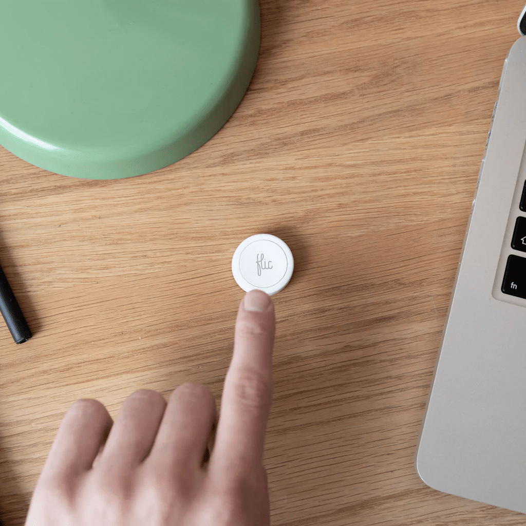 A hand pressing a Flic smart button on a wooden surface, with a laptop and green object in the background.