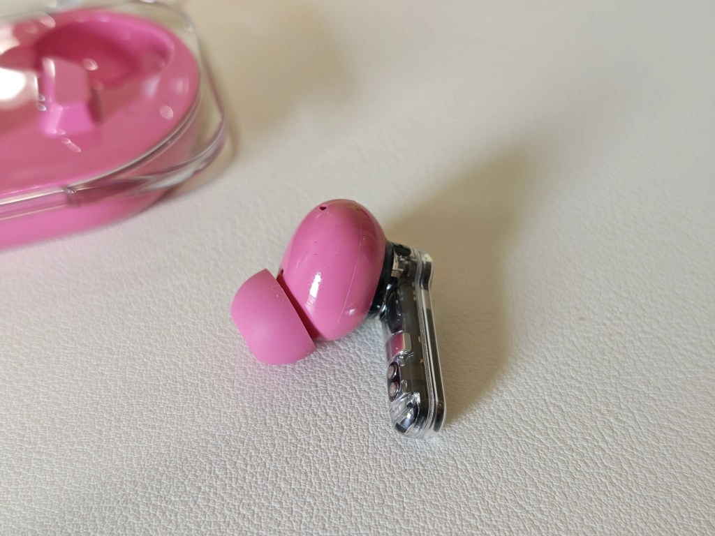 A close-up of a pink wireless earbud resting on a white textured surface, with a transparent charging case partially visible in the background.