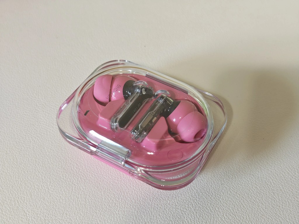 A pair of pink wireless earbuds resting in their transparent charging case on a light-colored surface.