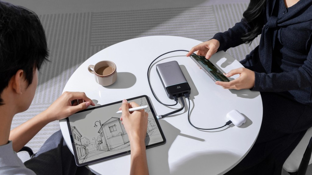 A person sketching on a tablet with a stylus while another person uses a smartphone, both seated at a table with a cup of coffee and a power bank connecting their devices.