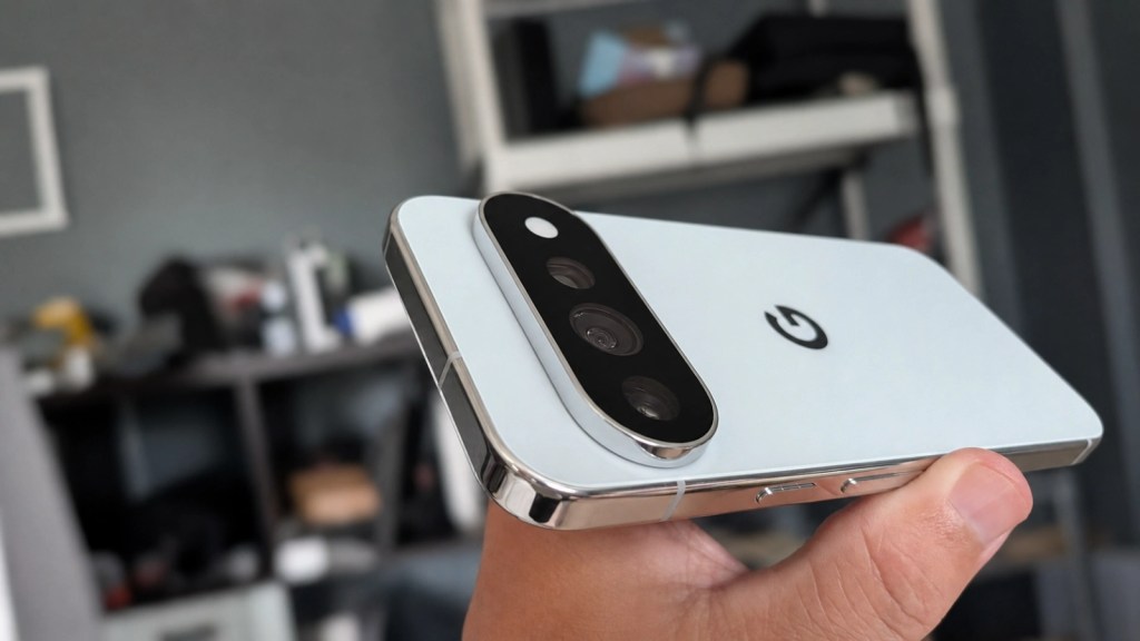 A person holding a smartphone with three rear cameras and a prominent logo on the back, against a blurred background of shelves.