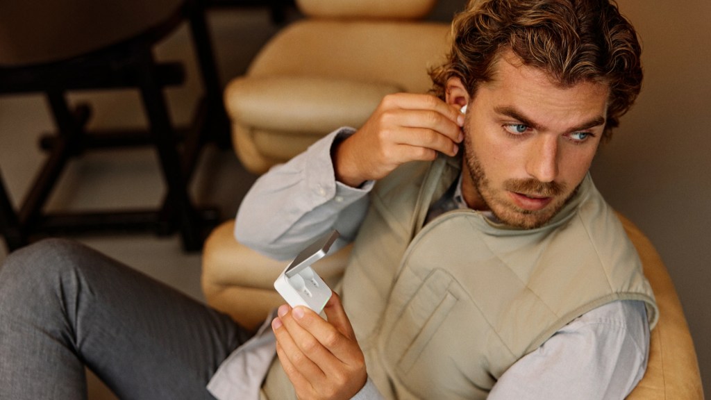 A man wearing a light vest is sitting in a chair, putting on wireless earbuds while holding a charging case.