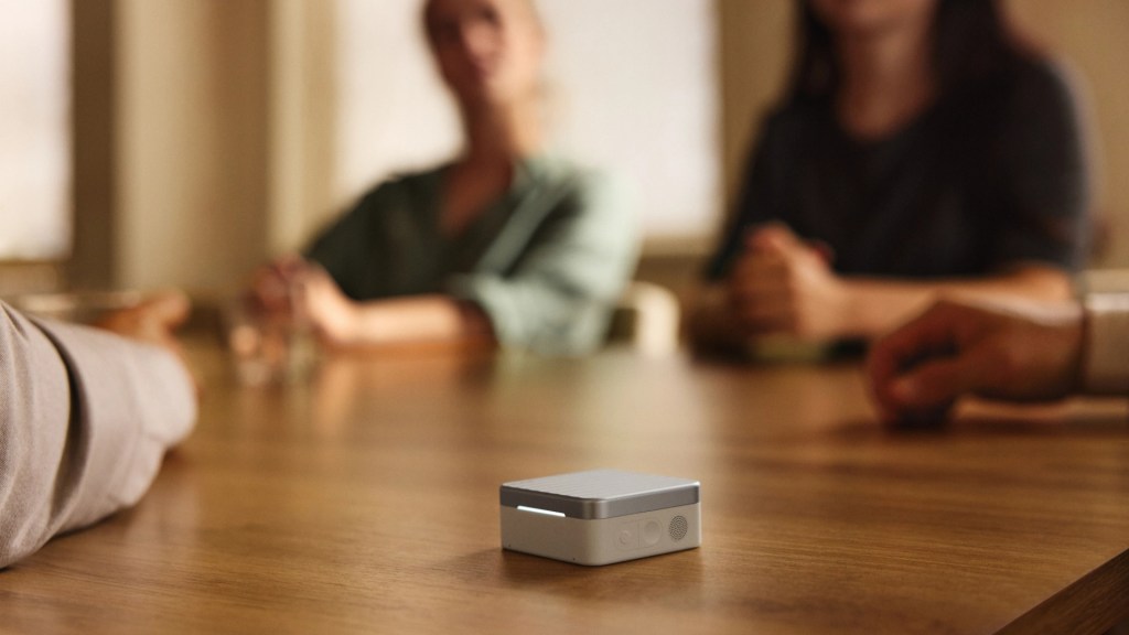 A close-up of a small, square device on a wooden table, with blurred figures of people sitting around the table in the background.