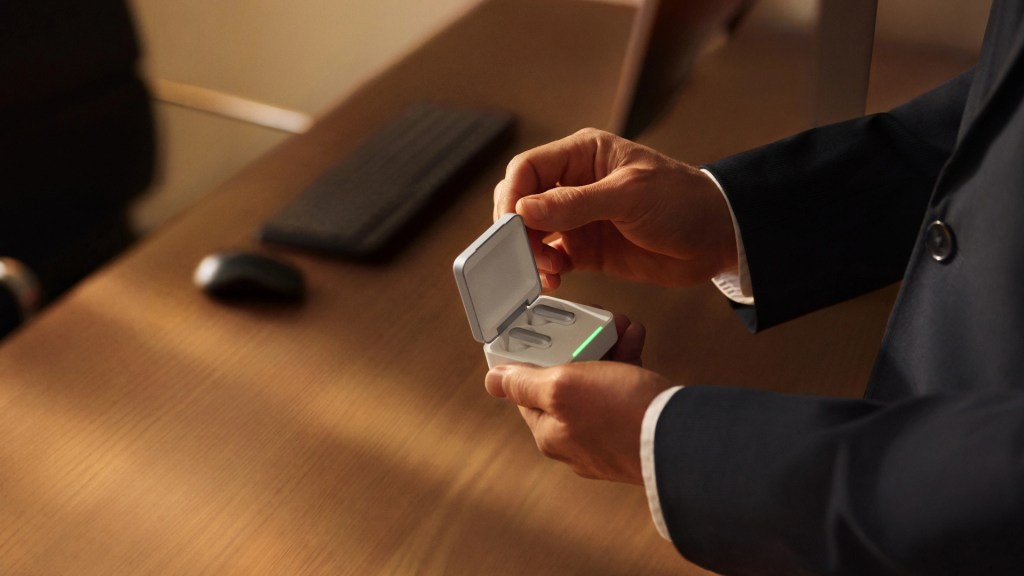 A person in a suit holding a wireless earbuds charging case with the lid open, displaying the earbuds inside, on a wooden desk.