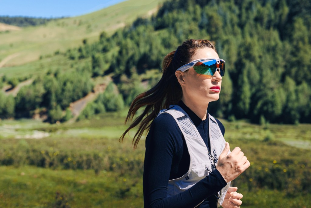 A woman running outdoors on a sunny day, wearing sports sunglasses and a lightweight vest, with green hills and trees in the background.