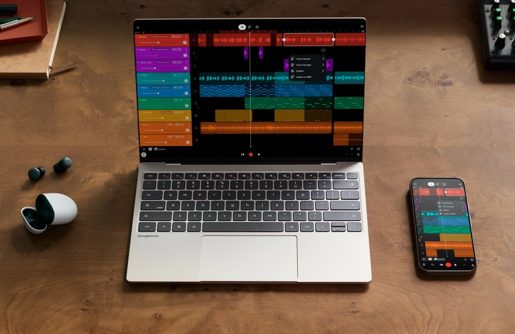 A laptop and smartphone displaying audio editing software with colorful waveforms on a wooden desk, alongside wireless earbuds and a notebook.