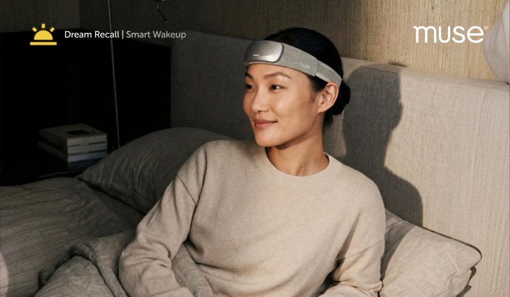 A woman sitting on a bed wearing a Muse headband, smiling as she engages with the Dream Recall Smart Wakeup feature.