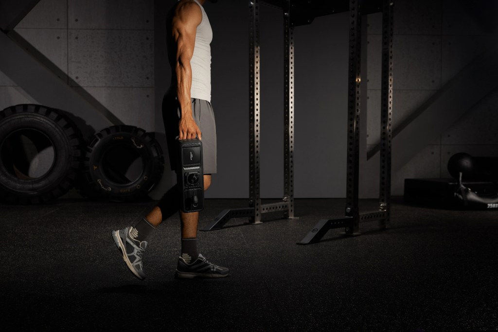 A person carrying a weight plate in a gym, with gym equipment and tires in the background.