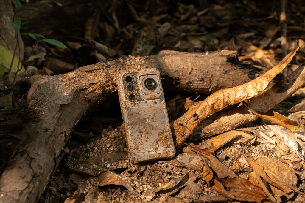 A smartphone lying partially buried in dirt and leaves on the forest floor.