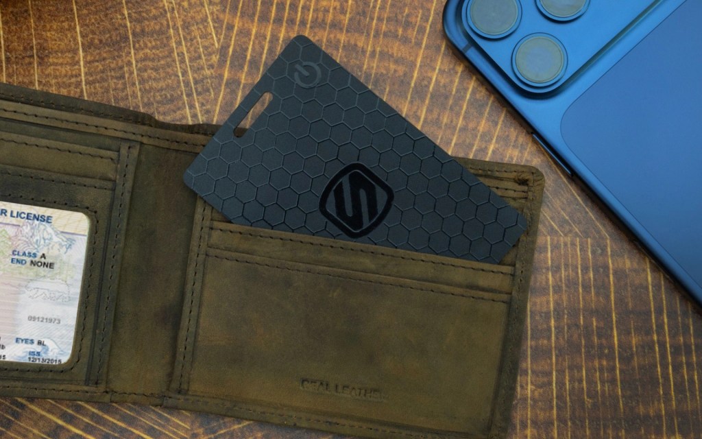 A close-up of a brown leather wallet opened to show a driver's license and a black hexagonal-patterned card, alongside a blue smartphone on a wooden surface.