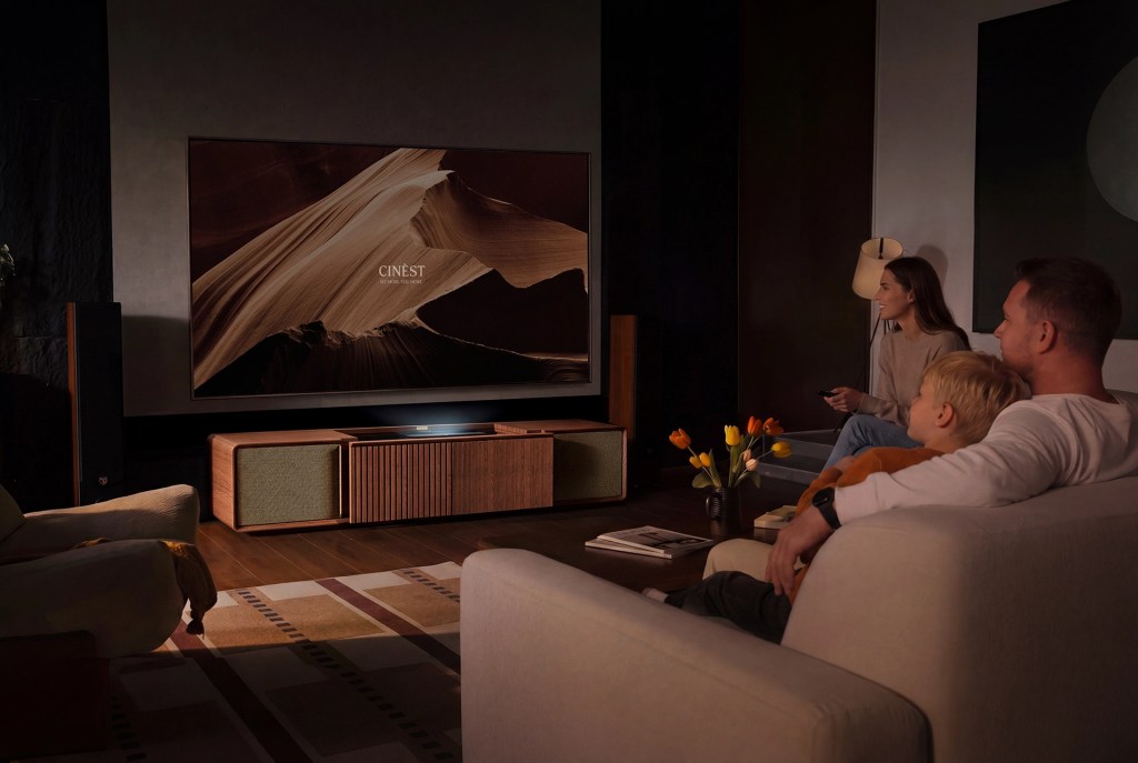 A family enjoying a movie night in a dimly lit living room, seated on a couch with a modern television displaying a cinematic image titled 'CINÉST'. A wood cabinet and a vase of tulips are visible.