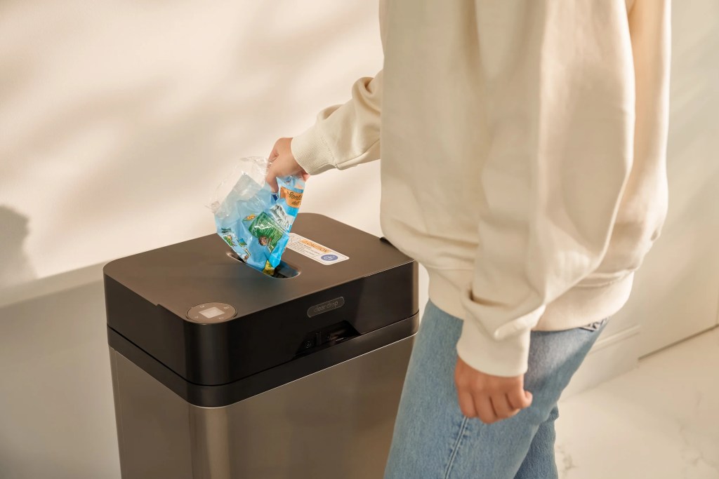 A person in a beige sweater disposing of a plastic bag containing items into a modern black trash can.