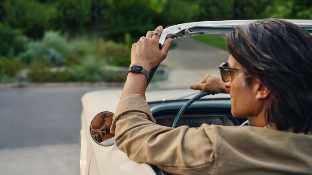 A man with long hair driving a convertible, resting his arm on the side of the car while wearing sunglasses and a watch.