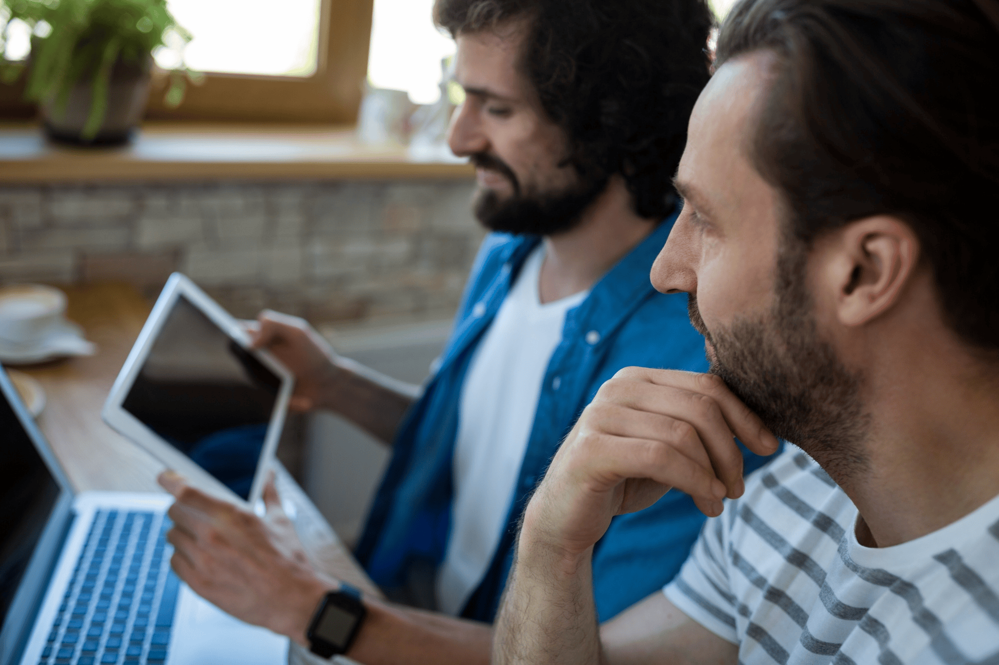 Two men focused on their devices while sitting at a wooden table, with one using a tablet and the other looking at a laptop. Bright indoor setting with natural light.