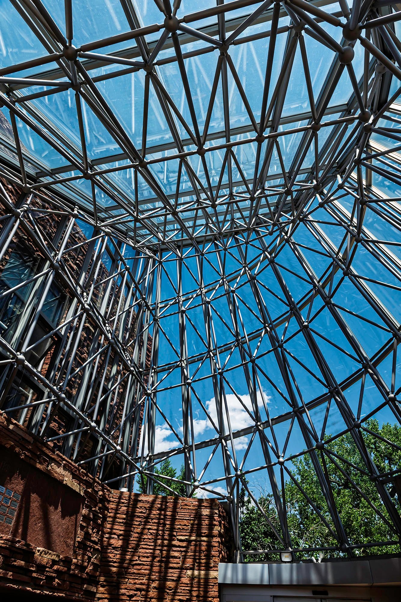Looking up through the steel and glass entrance pyramid of the Boulder Public Library, its triangular framing set against the Colorado sky above sandstone walls
