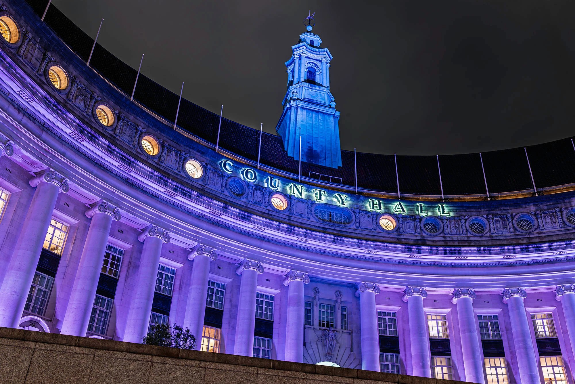 Low-angle night view of London County Hall on the South Bank illuminated in purple, showing the curved Edwardian Ionic colonnade, illuminated County Hall lettering on the parapet, and the central tower rising against a black London sky.