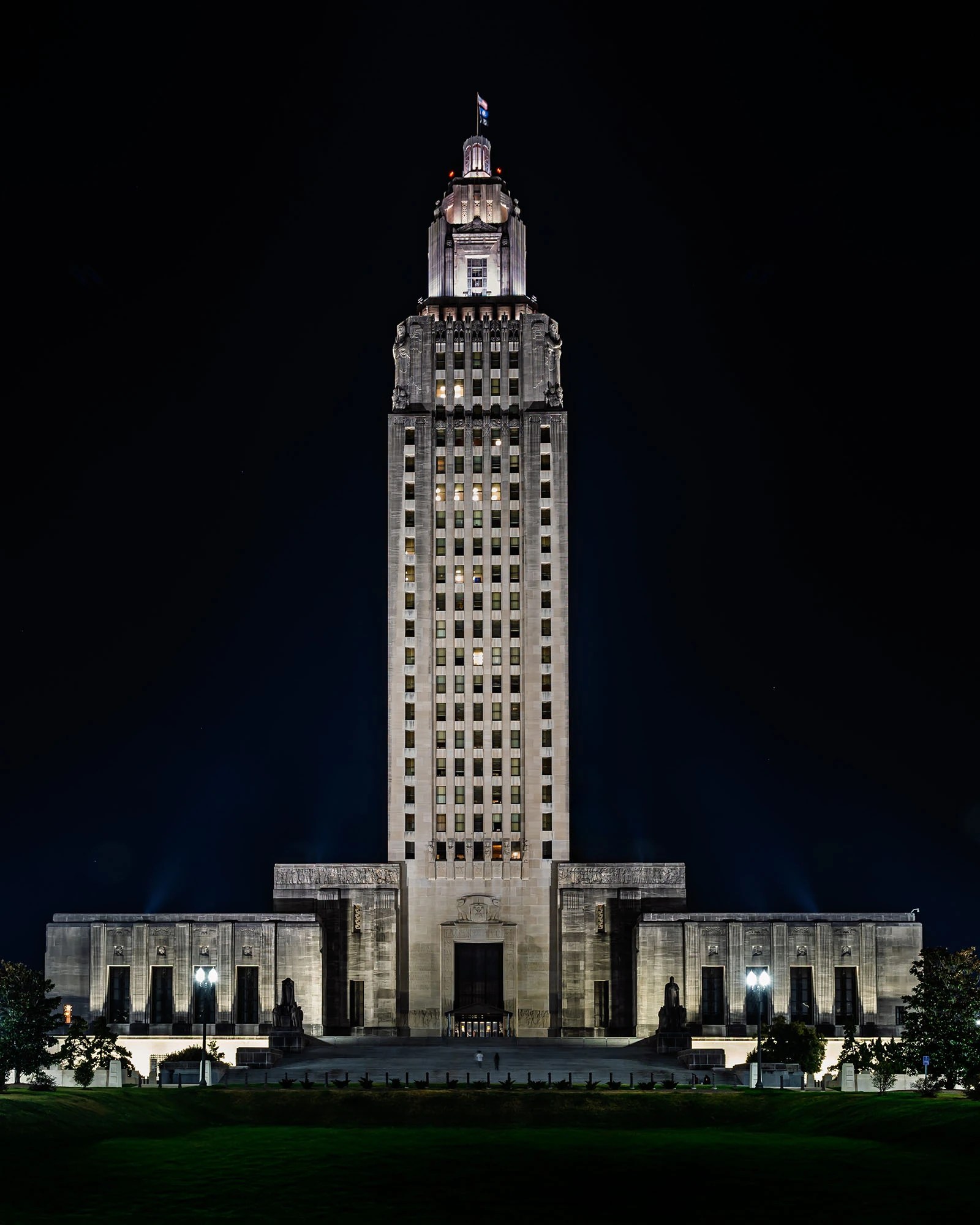 Full night view of the Louisiana State Capitol tower in Baton Rouge, showing the floodlit Art Deco limestone skyscraper rising from its monumental base against a black sky with the illuminated crown and Louisiana state flag at the top, green lawn in the foreground.