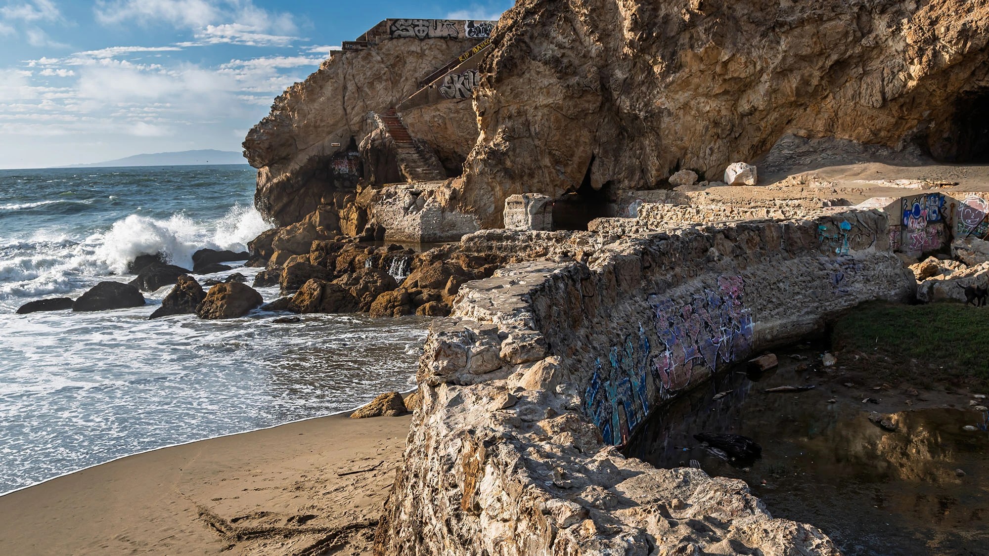 Ruins of the Sutro Baths at Land's End in San Francisco showing graffiti-covered concrete foundations, rocky cliffs with a metal staircase, sea caves, and Pacific Ocean waves crashing against the shoreline under a partly cloudy sky.