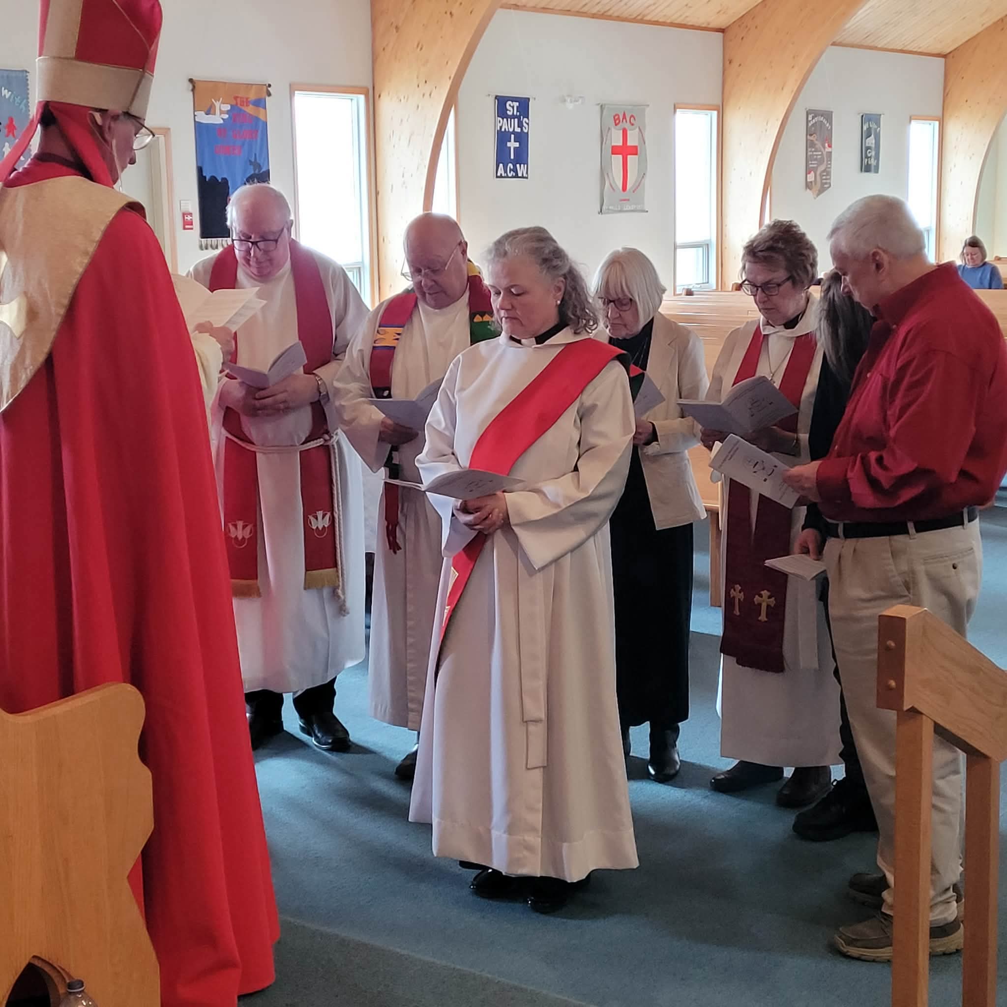 a woman wearing a deacon's red stole stands in front of a bishop. Behind her is a group of people