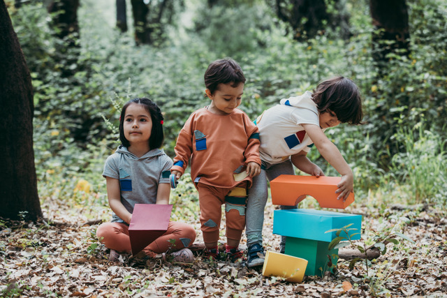 Três crianças, lado a lado, a brincar com blocos de construção, num bosque, e vestidas com as roupas da marca Miss Castelinhos - cool clothes, cool kids.