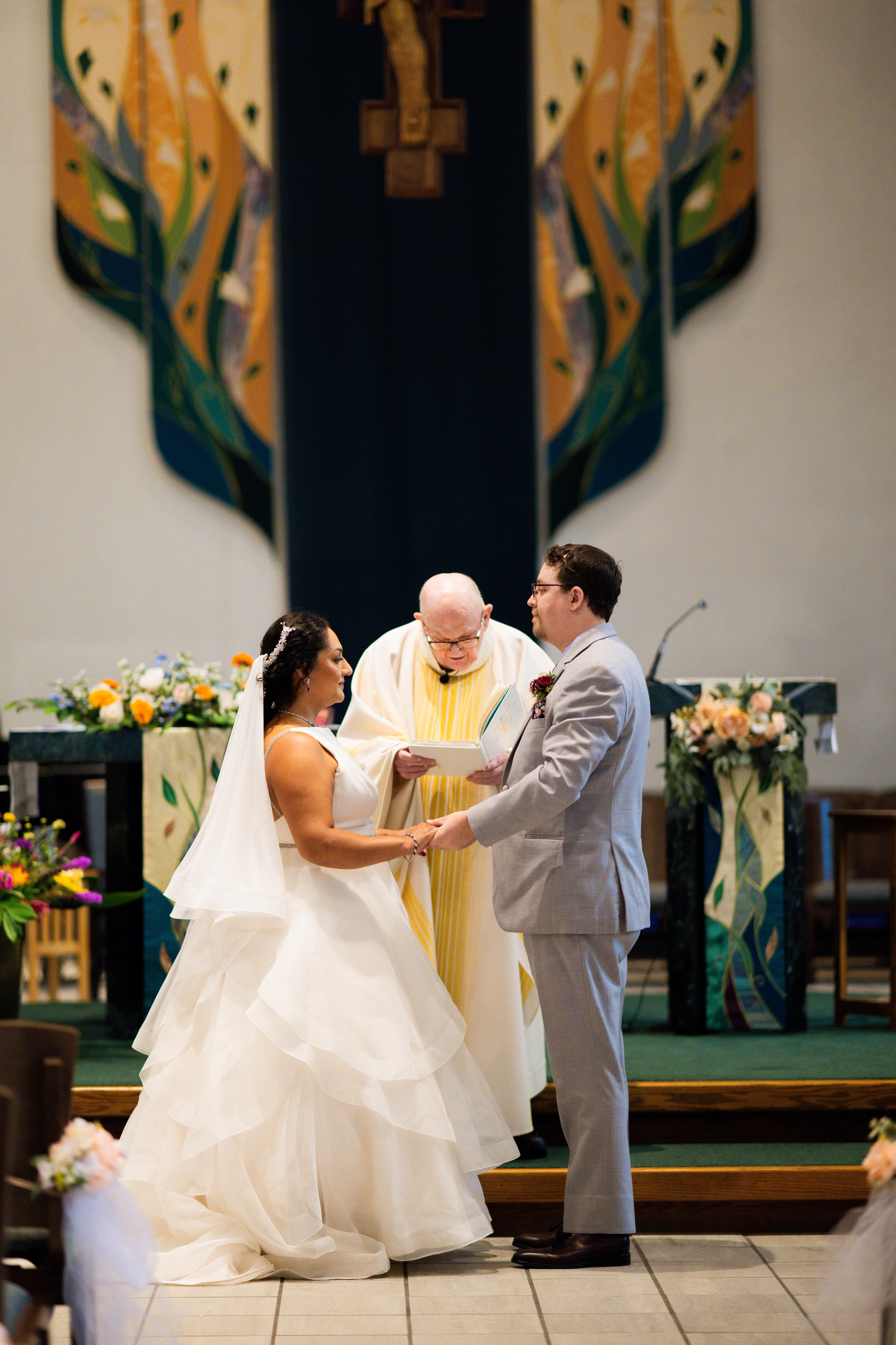 A bride and groom exchanging vows during a wedding ceremony, standing in front of a officiant in a decorated church.