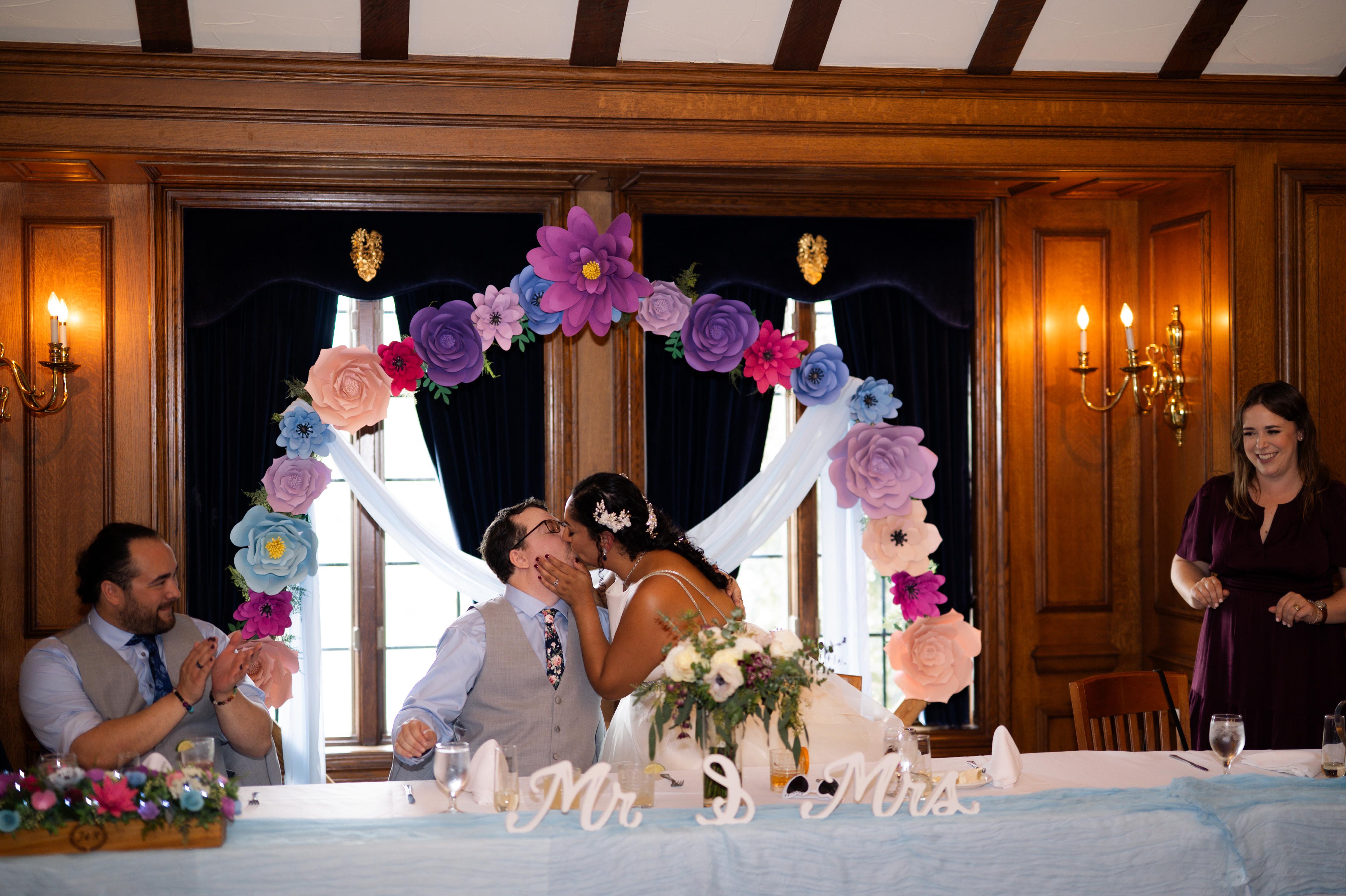 A newlywed couple shares a kiss during their wedding ceremony, framed by a decorative floral arch, while guests applaud and a woman smiles in the background.